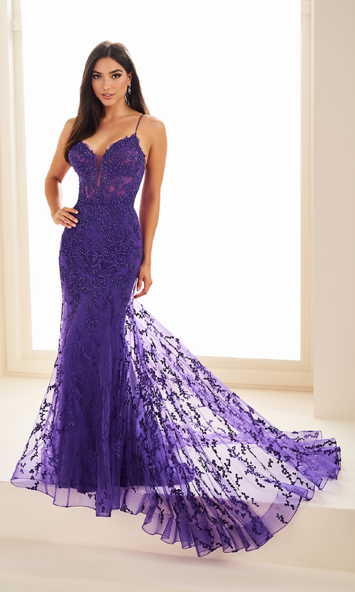A woman stands before a bright window in the Ellie Wilde EW36008—a fitted, floor-length purple mermaid prom dress with lace, sequin details, thin straps, and a flared semi-sheer train.